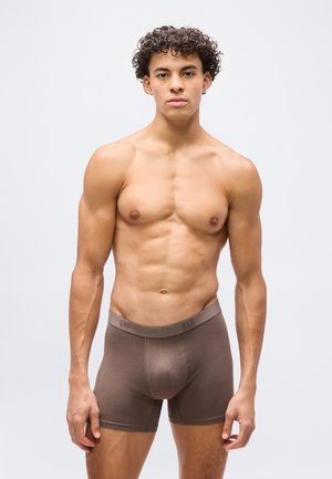 Νέος άνδρας με σγουρά μαλλιά, φορώντας στενά καφέ boxer, στέκεται μπροστά σε απλό ανοιχτό γκρι φόντο, κοιτάζοντας προς τα εμπρός.