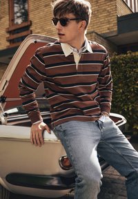 Brown and white striped long-sleeve polo with a collar, paired with light blue jeans. Model leans against a vintage white car.