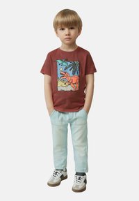 Rode katoenen T-shirt met een dinosaurusafbeelding, gecombineerd met lichtblauwe jeans en sneakers. Het shirt heeft een ronde hals en korte mouwen.
