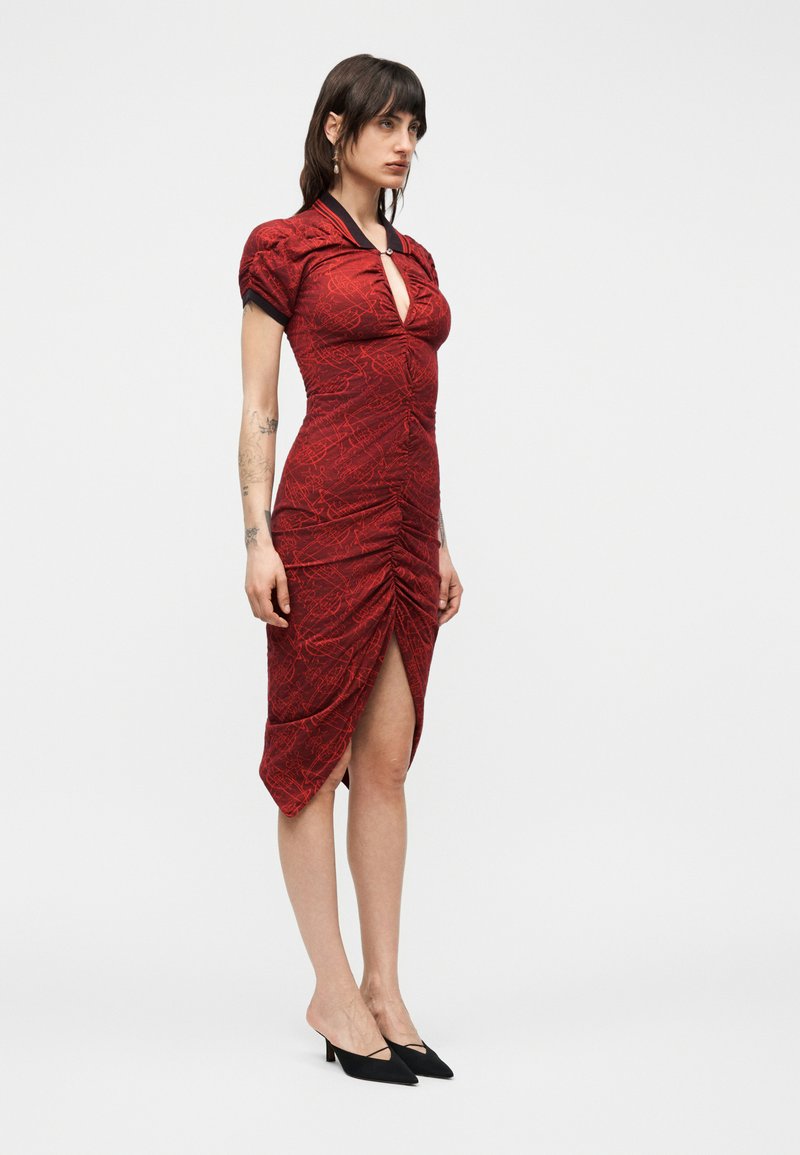 Woman stands wearing a fitted red ruched midi dress with a keyhole neckline, puff sleeves, black collar, side slit, and black low-heeled shoes.