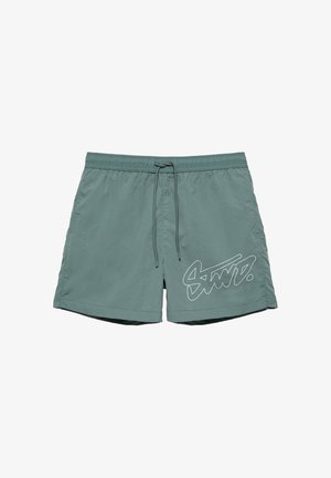 Short de bain vert avec taille élastique, cordon de serrage et logo blanc stylisé sur la partie inférieure de la jambe gauche.