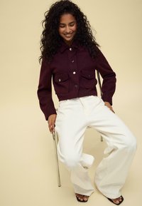 Maroon cropped jacket with two front pockets and silver buttons, paired with white wide-leg pants. Model seated on a silver chair.