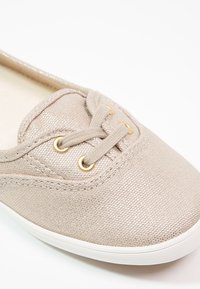 Baskets en toile beige avec une surface texturée, dotées d'œillets dorés et de lacets assortis. Semelle en caoutchouc blanche visible en bas.