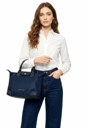 Jeune femme aux cheveux longs et ondulés portant une chemise blanche et un jean bleu, tenant un sac à main Valentino bleu marine avec des poignées en cuir noir.