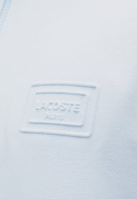 Tissu bleu clair avec un patch rectangulaire en relief présentant le nom de la marque "Lacoste Paris" en texte embossé.