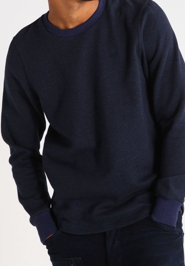 Homme portant un pull à manches longues bleu marine foncé avec poignets et col côtelés, mains partiellement dans les poches du pantalon.