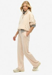 Vrouw in beige relaxte sweatshirt en bijpassende wijde broek met sneakers, staande met één hand in de zak tegen een witte achtergrond.