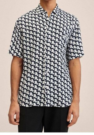 Mann trägt ein kurzärmeliges Hemd mit schwarz-weißem geometrischem Muster und schwarze Hose, steht vor einfachem Hintergrund.