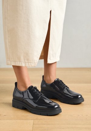 Scarpe in pelle verniciata nera con una spessa suola nera, caratterizzate da un design con lacci e punte tonde, indossate con una gonna beige chiaro.