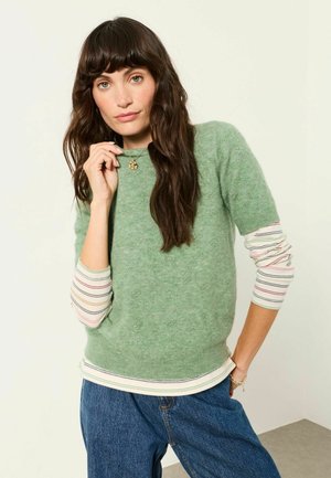 Frau mit langen dunklen Haaren, die einen grünen Pullover über einem gestreiften Hemd und blauen Jeans trägt, hält einen Halskettenanhänger nahe an ihrem Gesicht.