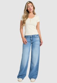 Top bianco a maniche corte con coste e tre bottoni anteriori, abbinato a jeans a gamba larga di colore azzurro chiaro. L'outfit presenta una vestibilità comoda e una texture liscia.