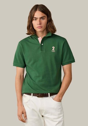 Jonge man met schouderlang bruin haar, die een groen poloshirt met nummer 2 en witte broek draagt, staat met één hand in zijn zak.