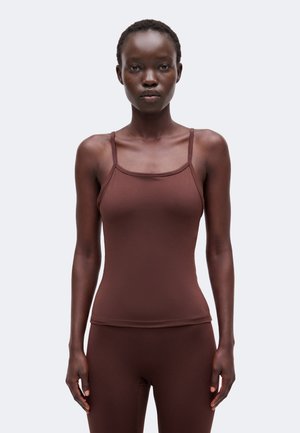 Femme aux cheveux courts portant un haut marron ajusté à fines bretelles et un bas assorti, debout devant un fond clair uni.