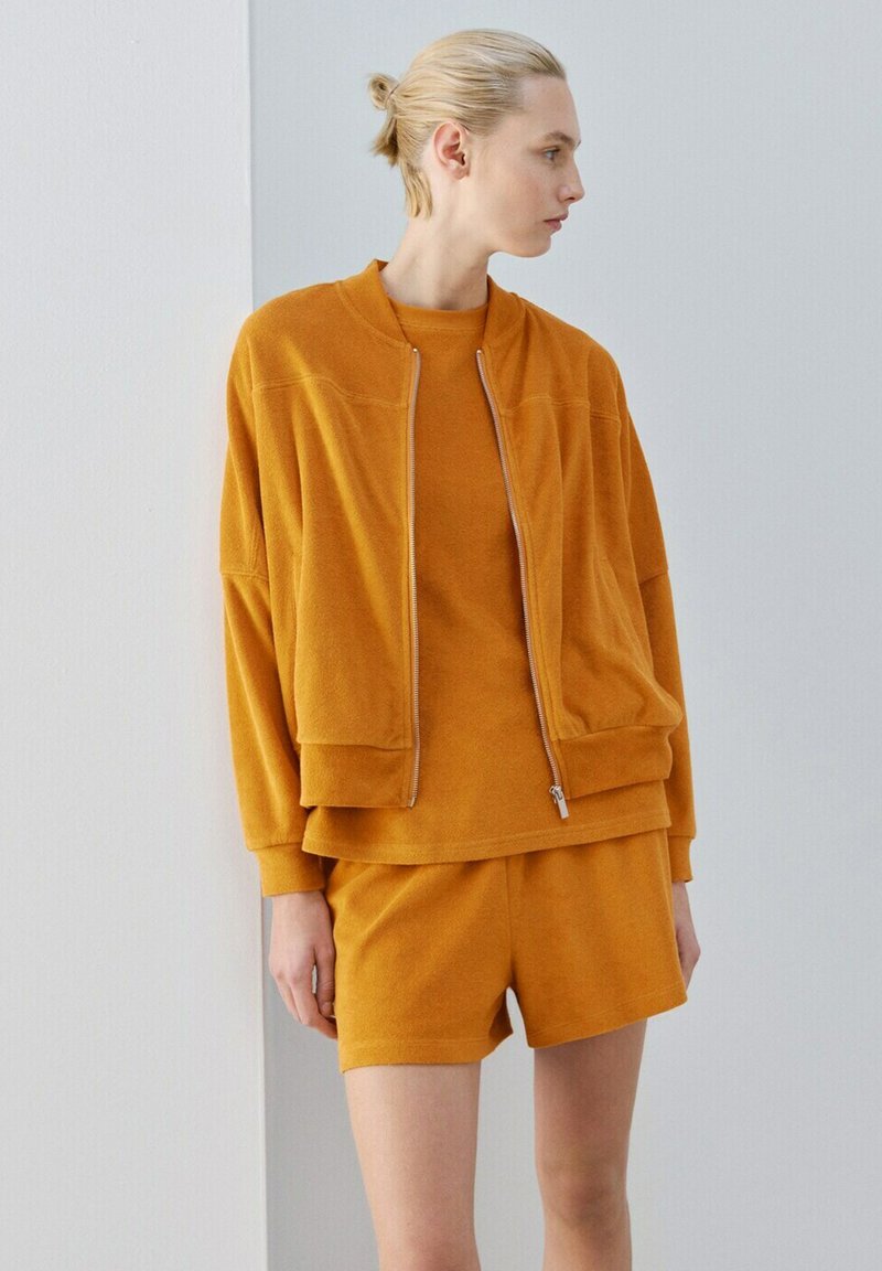 Femeie cu păr blond prins în coc poartă o jachetă portocalie cu fermoar, hanorac portocaliu asortat și pantaloni scurți portocalii, uitându-se spre dreapta pe un fundal simplu.