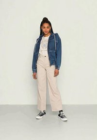 Casaco de ganga com cordões sobre uma t-shirt gráfica, combinado com calças de cintura alta em bege claro e sapatilhas negras.