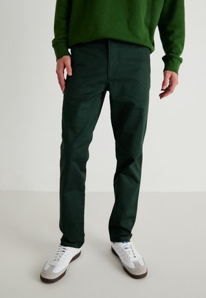 Pantalones de algodón verde oscuro con un corte de pierna recta. Combinados con zapatillas blancas con acentos en gris y azul. Textura suave, con detalles mínimos.