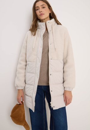 Veste rembourrée beige avec une partie supérieure douce et duveteuse et une partie inférieure lisse. Dispose d'un col montant, de cordons de serrage et de poignets élastiques.
