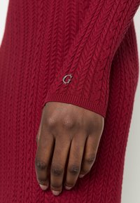 Punainen kaapelikudottu villapaita, jossa on ribbutettu pääntie ja hihansuut, sekä hopeinen logoyksityiskohta ranteen lähellä. Sujuva pinta ja slim-fit-muoti.