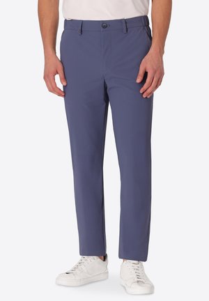 Uomo con pantaloni blu a gamba dritta e scarpe da ginnastica bianche, in piedi davanti a uno sfondo chiaro e uniforme.