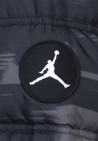 Detalle de una chaqueta de plumas negra con un parche redondo negro que presenta el logo blanco de Jumpman. Textura suave con un sutil patrón gris.