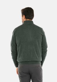 Maglione verde scuro con colletto alto, tessuto lavorato, design aderente e polsini a coste. Abbinato a pantaloni grigi con tasche posteriori.