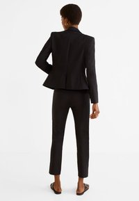 Upravený černý oblek s přiléhavým sakem s peplum detailem, úzkými kalhotami a nazouvacími sandály, vše v hladké, matné tkanině.