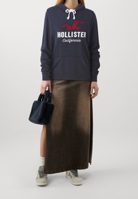 Marinblå hoodie med vit och röd "Hollister California"-logotyp, tillsammans med en brun satängkjol. Håller en marinblå plyschväska. Grå sneakers med vita accenter.