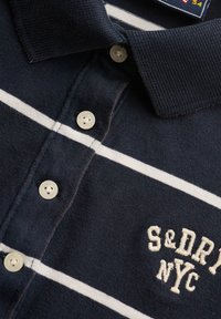 Polo blu navy con colletto, righe orizzontali bianche, bottoni bianchi e testo ricamato "S&DRJ NYC" sul davanti.