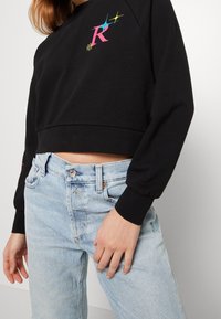 Felpa nera corta con un grafico colorato della "R" e una stella. Jeans in denim blu con vestibilità classica e lievi segnali di usura sui taschini.