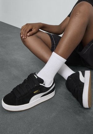 Baskets en suède noir avec des accents blancs et de grosses lacets noirs. Elles disposent d'une semelle en caoutchouc texturée et d'un détail en logo doré sur le côté.