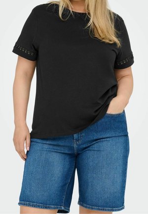 Frau trägt ein schwarzes Kurzarm-Top mit Spitzenbesatz und blaue Jeans-Shorts bis zum Knie, steht mit einer Hand in der Tasche.