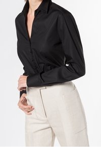 Donna che indossa una camicia nera aderente con bottoni infilata in pantaloni beige a vita alta con grandi tasche laterali.