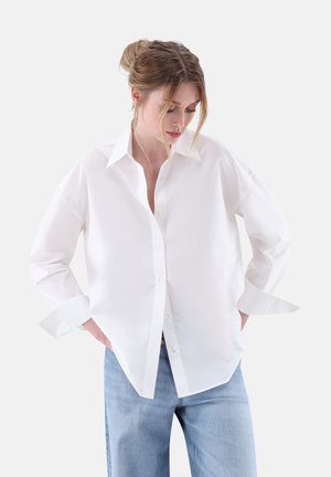 Camicia bianca con colletto a revers, maniche lunghe e dettagli ai polsini, realizzata in tessuto leggero, abbinata a jeans azzurri.