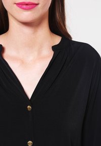 Černá blůza s plisovaným výstřihem, zlatými knoflíkovými detaily a hladkou, lehkou texturou, která zdůrazňuje jednoduchý a elegantní design.