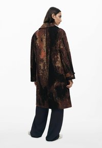 Long manteau texturé noir avec des motifs abstraits orange et dorés. Présente un large col et des épaules tombantes, associé à un pantalon sombre.