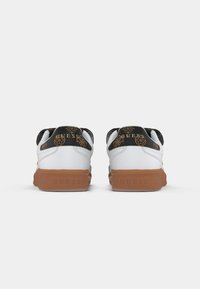 Fehér sportcipő fekete mintás hátrésszel, arany logóelemekkel és barna gumi talppal. Texturált részletek a saroknál.