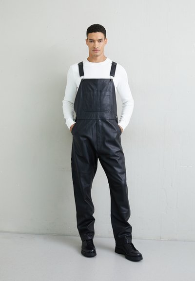 Zwarte leren overalls hebben verstelbare banden, een voorzak en een losse pasvorm. Het model combineert ze met een wit overhemd met lange mouwen en zwarte schoenen.