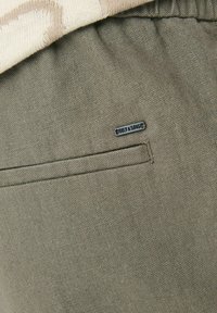 Khaki linnedragter med en subtil tekstur, der har en baglomme og et lille mærke i metalaccent.