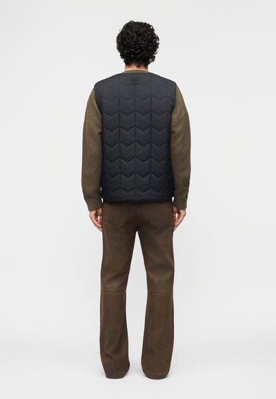 Άνδρας με σγουρά μαλλιά, που φοράει μαύρο γιλέκο με καπιτονέ σχέδιο πάνω από καφέ υφασμάτινο πουλόβερ και καφέ παντελόνι, στέκεται με την πλάτη προς τα εμάς σε λευκό φόντο.