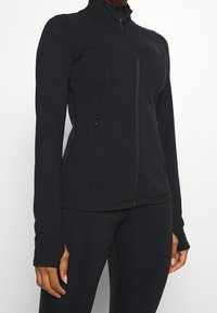 Černá zipová bunda s dlouhými rukávy, vyrobená z elastického materiálu. Obsahuje boční kapsy a vysoký límec se švy.