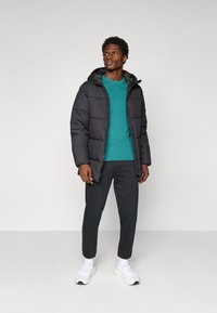 Giacca a piumino nera con cappuccio, felpa teal sotto, pantaloni scuri e sneakers bianche. Tessuto liscio con design trapuntato.