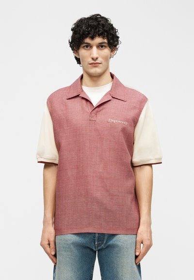 Jeune homme aux cheveux bouclés et foncés portant une chemise à manches courtes rouge à carreaux par-dessus un t-shirt crème et un jean bleu, faisant face à l'avant.