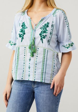 Lyseblå bluse med korte ærmer, grøn blomsterbroderi, v-udskæring og to grønne kvaster ved halsudskæringen. Har en afslappet pasform.