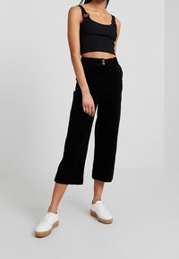 Pantalons en velours côtelé noirs, coupés et à jambe large, associés à un débardeur noir ajusté. La tenue est complétée par des baskets à plateforme blanches.