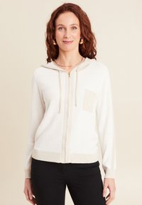 Cardigan à capuche crème avec fermeture éclair, tricot texturé, poignets côtelés et une petite poche en patch d'une teinte plus claire de crème.