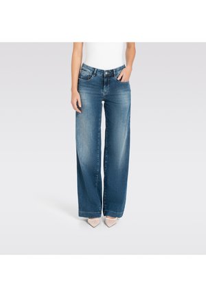 Frau trägt hoch taillierte, weit geschnittene blaue Jeans mit Fronttaschen, kombiniert mit weißen, spitz zulaufenden Absätzen, vor einem hellgrauen Hintergrund stehend.