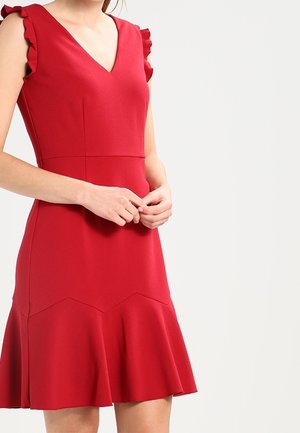 Nainen pukeutuneena hihattomaan punaiseen mekkoon, jossa on röyhelöiset olkapäät, V-muotoinen pääntie, vyötärö istuu tiukasti ja helma levenee, seisoo vaaleaa yksinkertaista taustaa vasten.