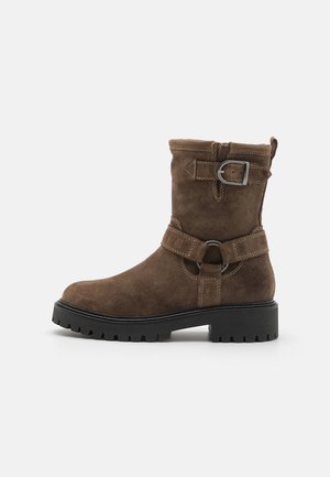 Botte mi-mollet en daim marron avec semelle à crampons noire, sangle décorative à boucle près du sommet et sangle de style harnais avec anneau en métal autour de la cheville.