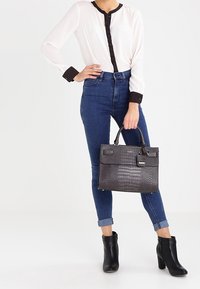 Kobieta w białej bluzce z czarnymi wykończeniami, niebieskich, podwiniętych rurkach, czarnych botkach za kostkę, trzymająca torebkę z teksturowanej czerni.