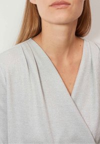 Světle šedý top s překryvem, který má jemný lesk a texturovanou tkaninu, s draped záhyby a hlubokým výstřihem do V.